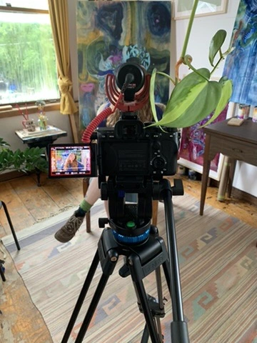 A behind the scenes photo of a camera pointed at a woman giving an interview